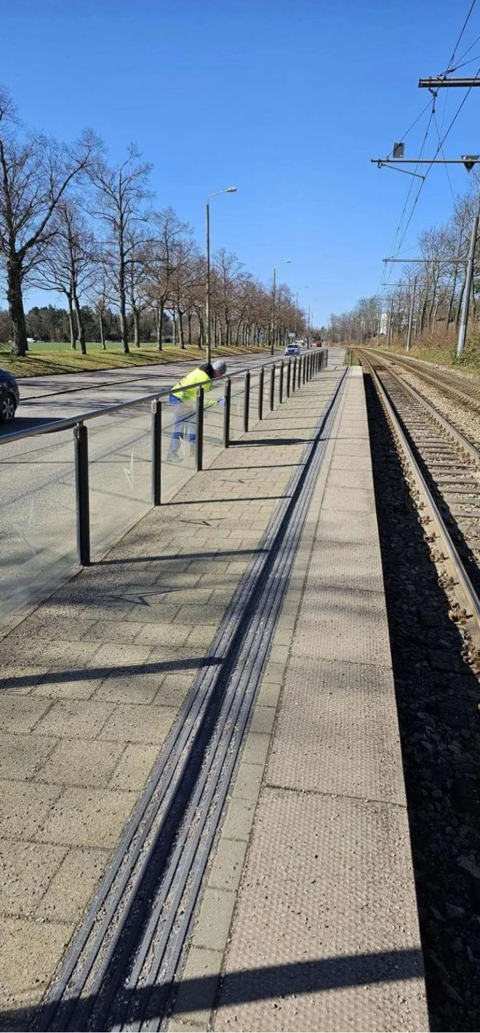 Moderne Raumpflege GmbH Boge - Gebäudereinigung – eine Reinigungskraft reinigt eine Sicherheitsbarriere aus Glas und Metall entlang der Gleise